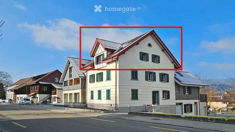 A two-story residential building with a white facade, green shutters, and a red-tiled roof. The house features multiple windows and a small balcony on the upper floor.