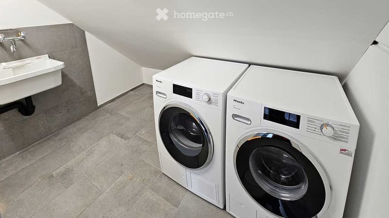 Modern bathroom with a washing machine, dryer, and a small sink, featuring tiled flooring and white walls.