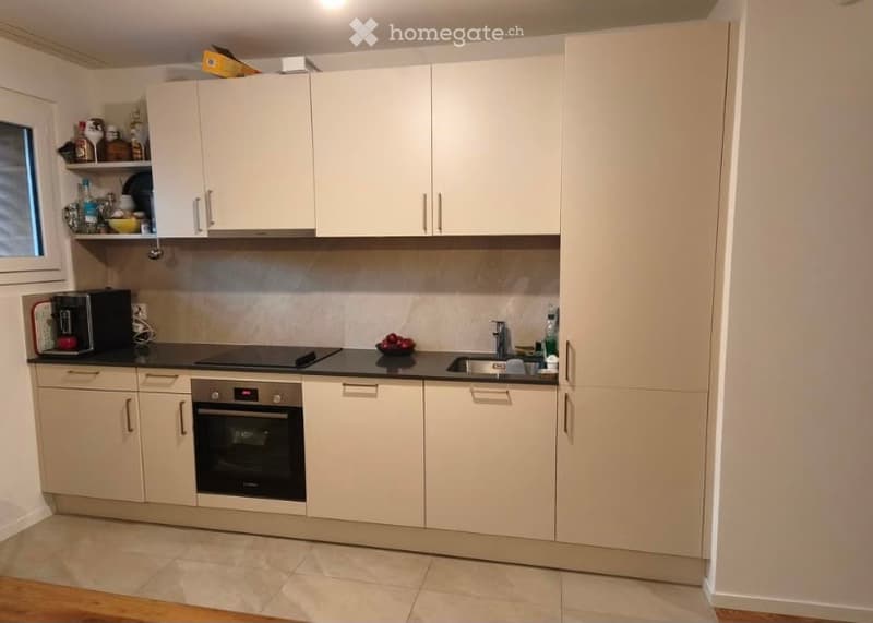 Modern kitchen with white cabinets, black countertops, and stainless steel appliances including an oven, sink, and coffee maker. A small window is visible on the left side.