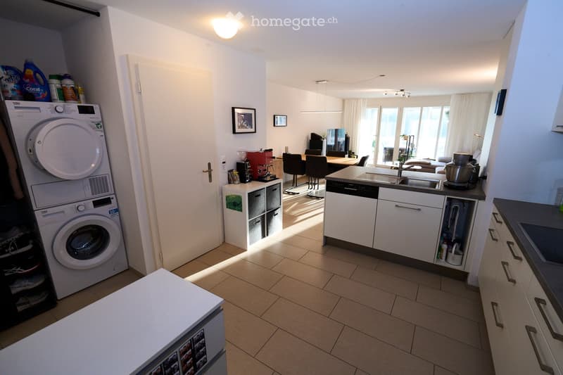 Open-plan kitchen and living area with white cabinets, a central island, a washing machine, and a dining area with chairs and a table.