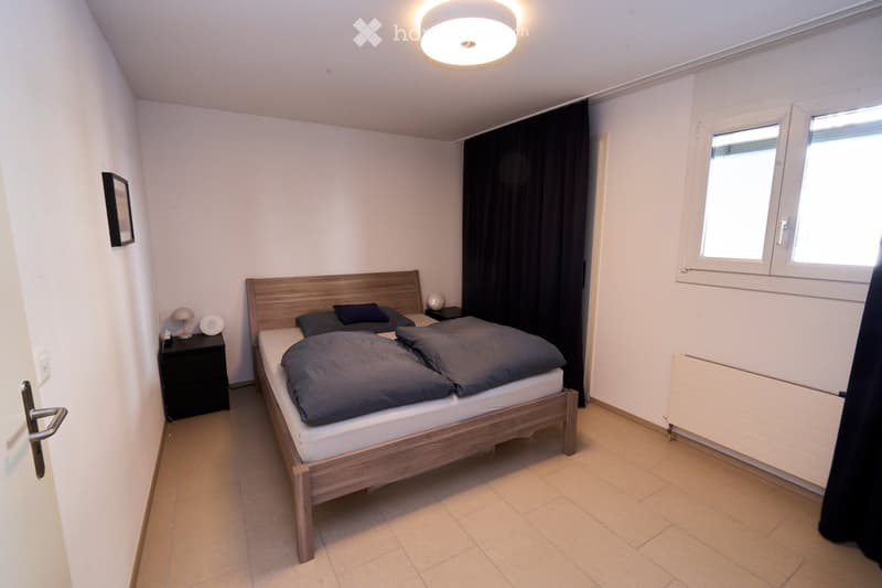 Cozy bedroom with a wooden bed, nightstand, lamp, and picture frame on the wall. The room features tiled flooring and large windows with dark curtains.