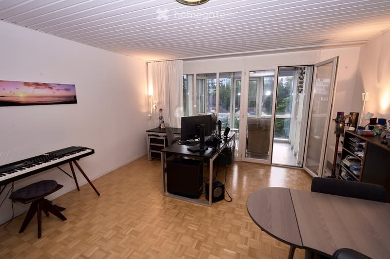 A well-lit room with wooden flooring, a desk with a computer setup, a keyboard on a stand, a round table with chairs, and a shelf with books and decorative items. Large windows and sliding glass doors lead to a balcony area.