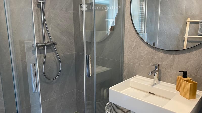white tiles, glass shower enclosure, round mirror, modern faucet, white vanity with sink, soap dispenser, shelves with toiletries