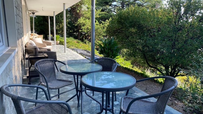 balcony with tables and chairs, covered with ceiling, stone wall, floor paved with tiles, trees and greenery, mountain view