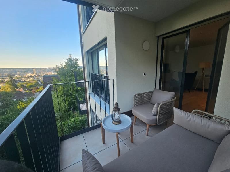Balcony with a view of the city, featuring a sofa, a chair, a small table with a lantern, and a railing.