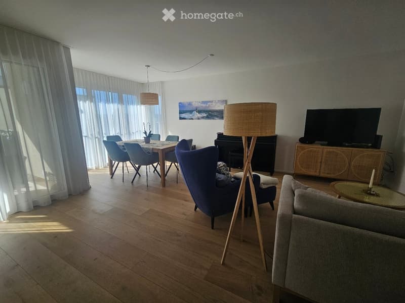 Bright living room with wooden flooring, large windows with white curtains, a sofa, chairs, a dining table, a television, and a lamp.