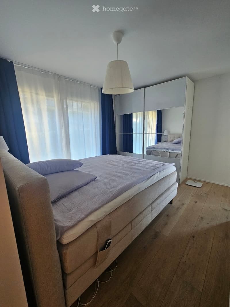 Modern bedroom with a large bed, mirrored wardrobe, pendant light, and sheer curtains allowing natural light.