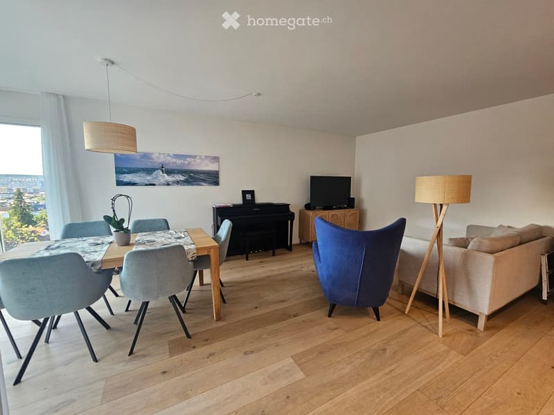 Modern living and dining area with light wooden flooring, a sofa, a dining table with chairs, a piano, a television, and a potted plant.