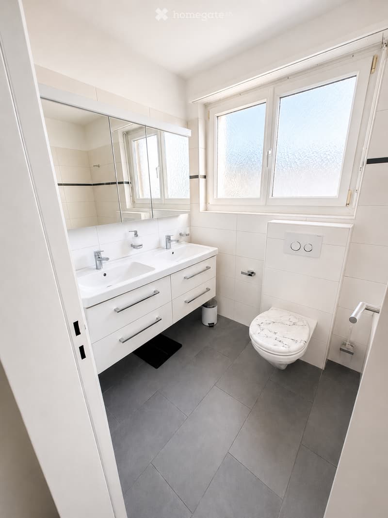 Modern bathroom with white tiles, a double sink, a large mirror, and a marble toilet seat. The room features grey tiled flooring and large windows with frosted glass.
