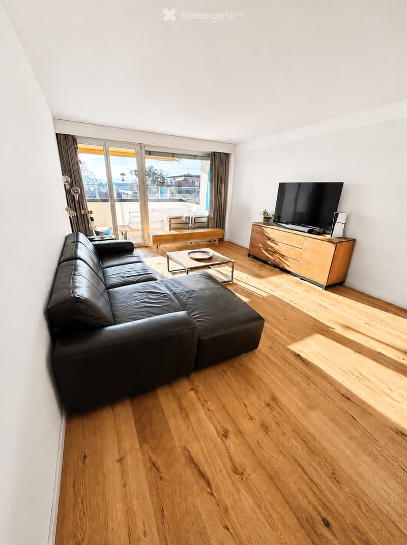 Modern living room with wooden flooring, large sliding glass doors leading to a balcony, a black leather sofa, a coffee table, a TV stand, and a small table with chairs.