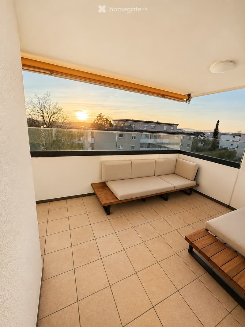 Balcony with tiled floor, white walls, large window offering a view of the sunset, and a cream-colored sofa with wooden tables.