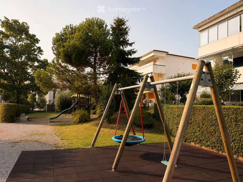 Playground area with swings, slide, and trees, surrounded by modern residential buildings.