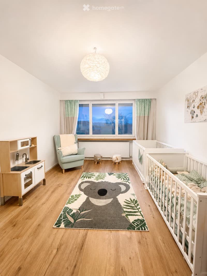 Bright, modern children's bedroom with wooden flooring, large window with green curtains, baby crib, play kitchen, cozy armchair, decorative rug with koala design, and two small animal-shaped stools.