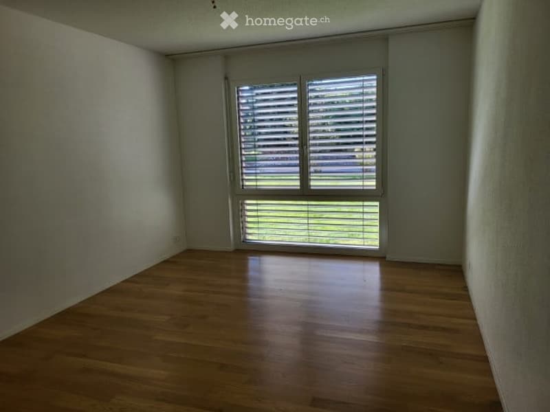 Empty room with wooden flooring and large window with blinds, showing a view of greenery outside.