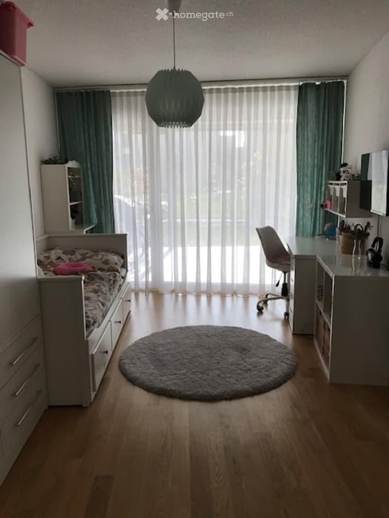 A modern, well-lit bedroom with a bed, desk, chair, wardrobe, and large window with curtains. The room features a round rug and a stylish hanging lamp.
