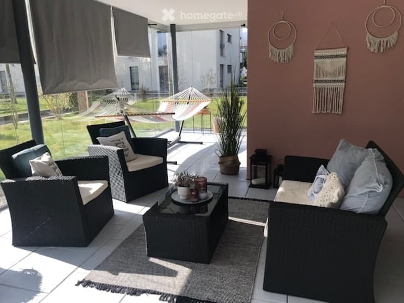 A modern, furnished balcony or terrace with wicker chairs, a coffee table, and decorative elements like dreamcatchers and lanterns.