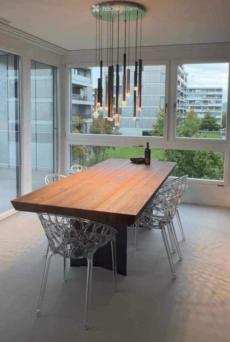A dining room with a large wooden dining table, four transparent chairs on each side, and a large glass door leading to a view outside
