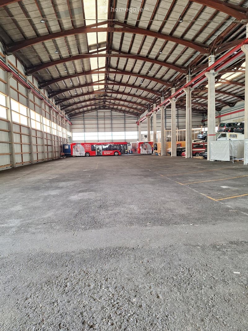 Large industrial-style building with a high, metal-framed roof and concrete floor, featuring several parked vehicles including buses and vans along the sides.