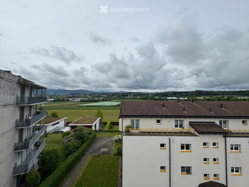 A multi-story residential building with balconies and surrounding greenery, set against a rural landscape with fields and distant hills.