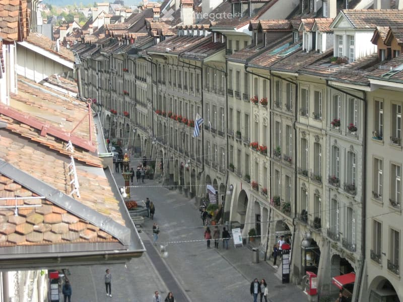 A historic European street lined with multi-story buildings featuring red-tiled roofs, ornate facades, and flower boxes. People walk along the cobblestone street, and flags are visible hanging between buildings.