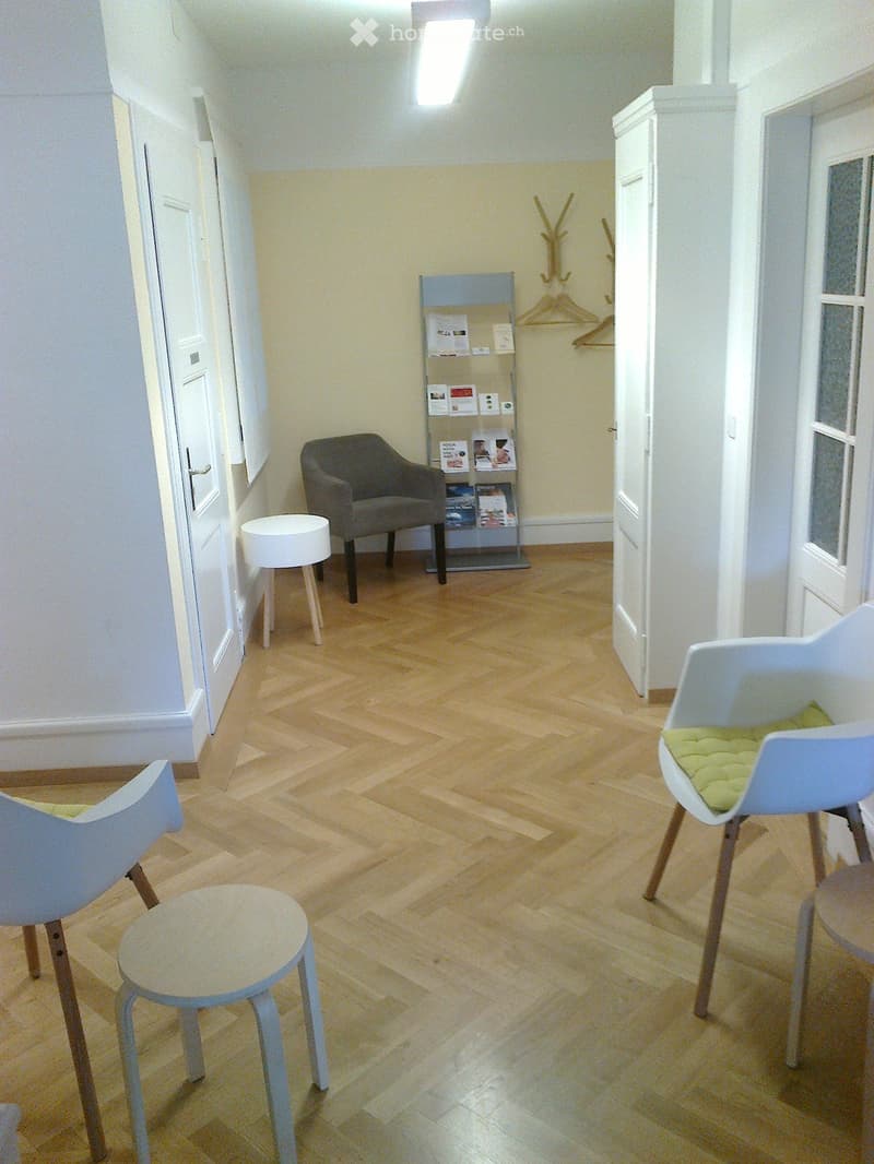 A small, well-lit room with wooden flooring, featuring a grey armchair, two white stools, and a small shelf with brochures.