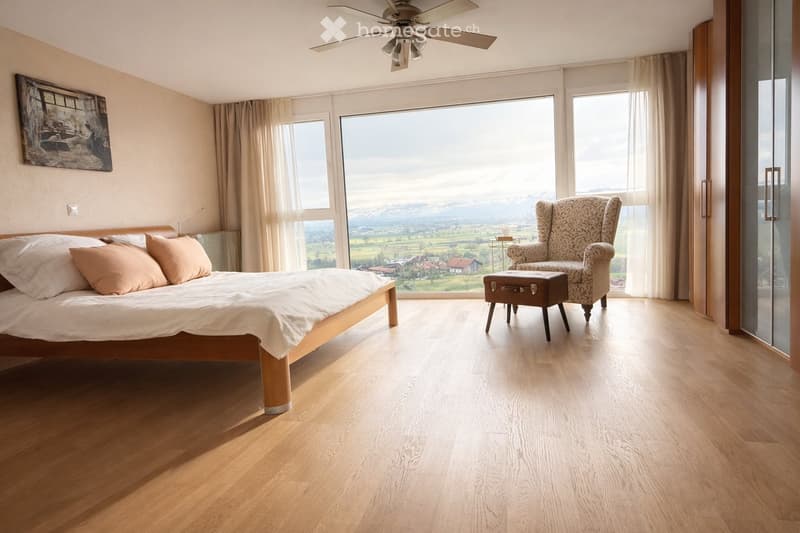 Spacious bedroom with wooden flooring, large windows offering a scenic view, a bed with pillows and a blanket, a patterned armchair, a small stool, and a picture frame on the wall.