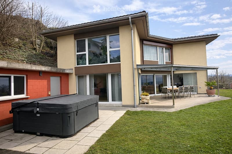 Modern two-story house with large windows, a covered patio area with a dining set, a hot tub, and a well-maintained lawn.