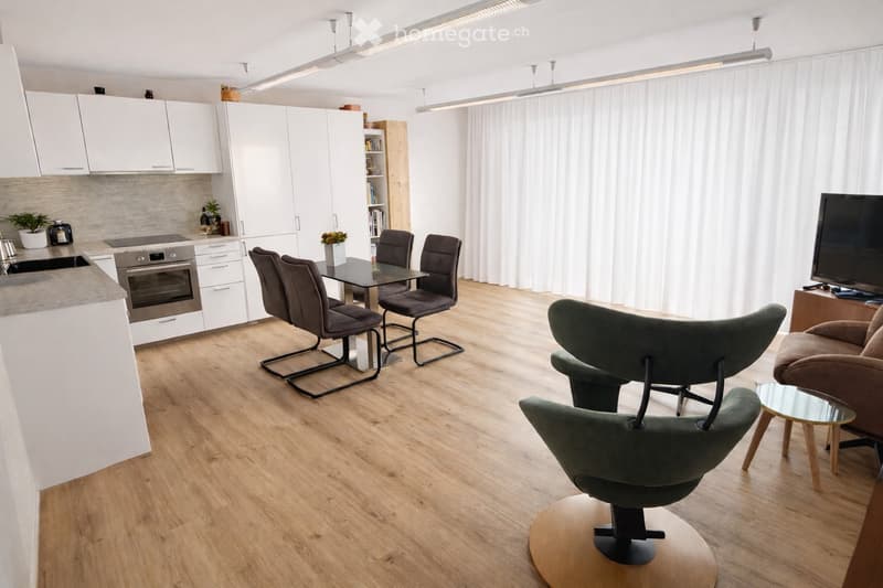 Open-plan living and dining area with modern furniture, including a sofa, chairs, a dining table, and an integrated kitchen with white cabinets and stainless steel appliances.