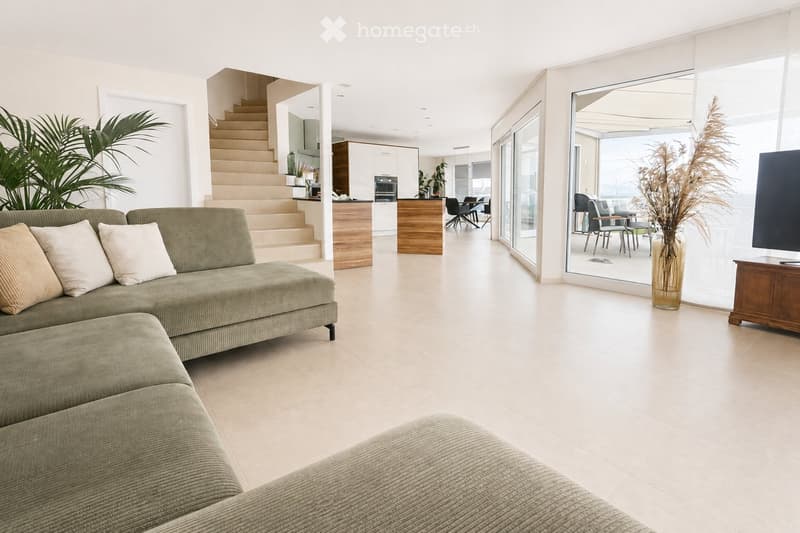 Modern living room with a green sectional sofa, coffee table, dining area with chairs and table, large glass doors leading to a balcony, and decorative plants.