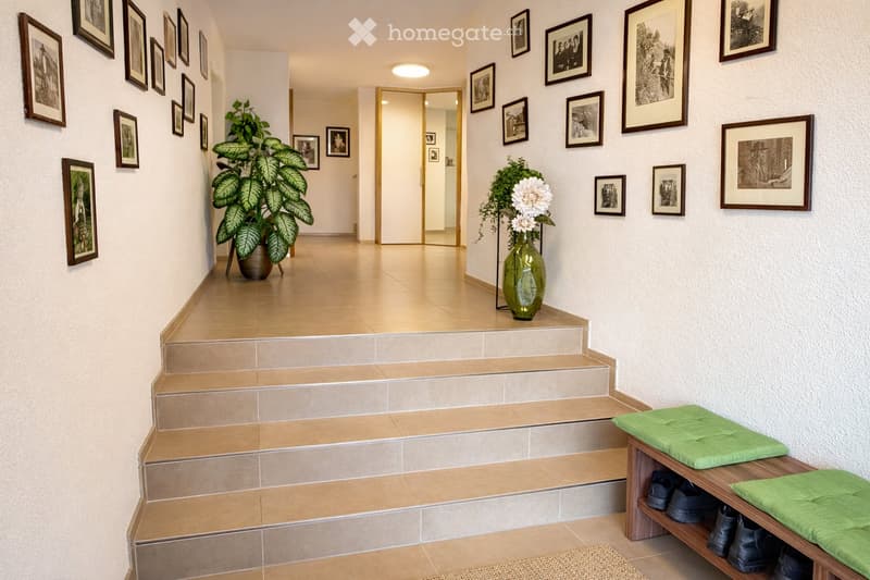Hallway with tiled floor, framed pictures on walls, green cushioned bench, potted plants, and a staircase leading upwards.