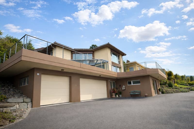 Two-story modern house with beige and brown exterior, large garage, balcony with glass railing, and landscaped garden area.