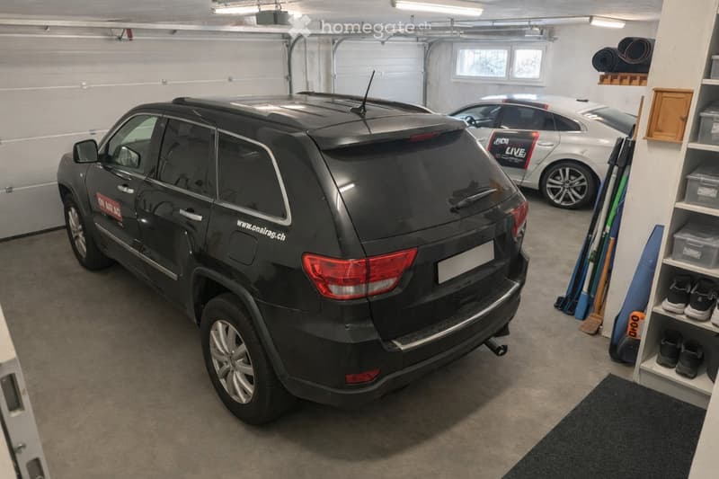 Garage with two parked cars, one black SUV and one silver sedan, along with storage shelves and cleaning tools.