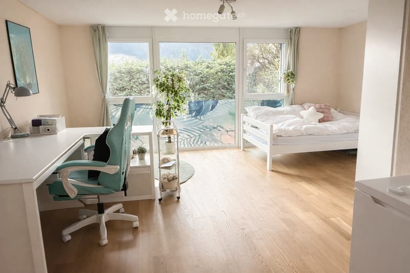 A modern, well-lit room with a desk and chair on the left, a bed on the right, and large windows with a view of greenery. The room features a light wooden floor, a potted plant, and a small shelf with books and decorative items.