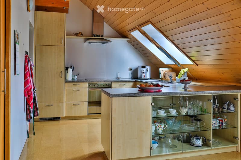 Bright kitchen with wooden cabinets, stainless steel appliances, and a central island with glass-fronted storage. The space features a skylight, a coffee maker, and a bowl of fruit on the counter.