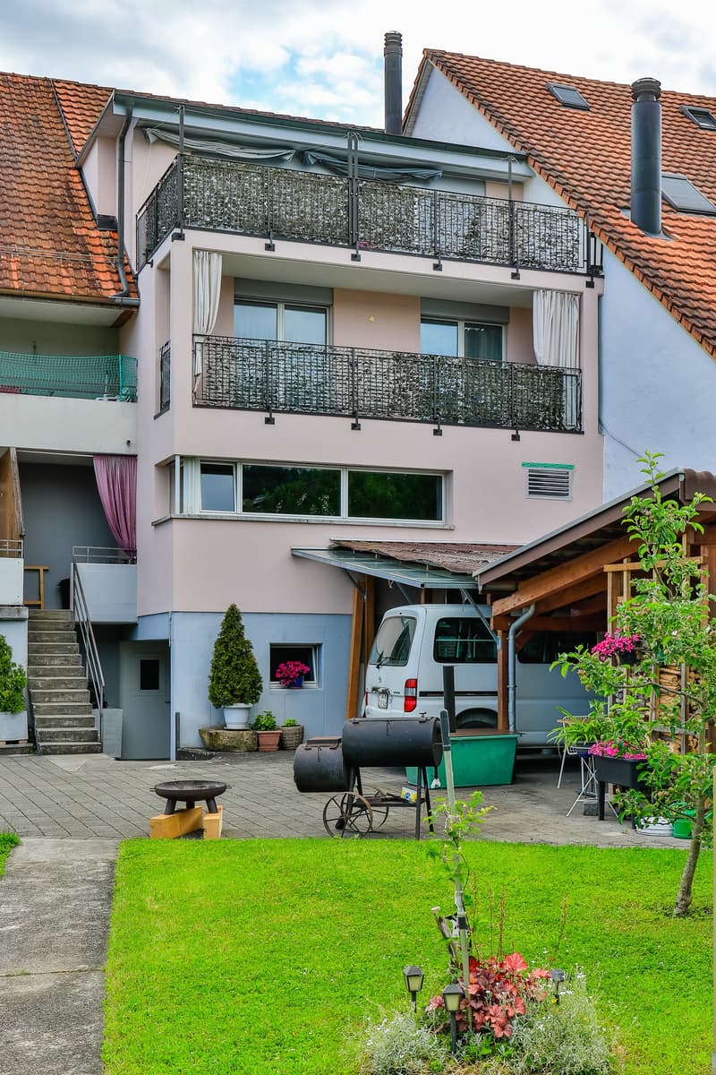 A two-story house with a red-tiled roof, balconies, and a well-maintained garden featuring a lawn, flower beds, and outdoor furniture.