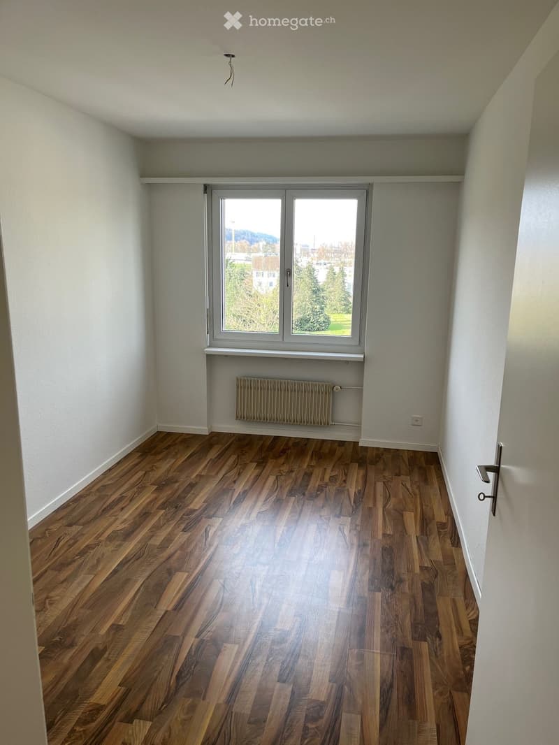 Empty room with wooden flooring, white walls, large window with view, radiator, and door handle visible.