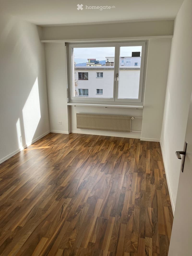 Empty room with wooden flooring, white walls, large window with view, radiator, and door.