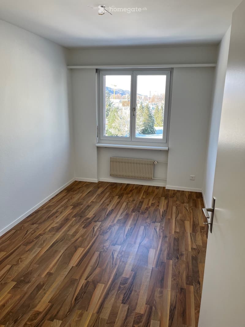 Empty room with wooden flooring, white walls, large window with view, radiator, and door handle visible.