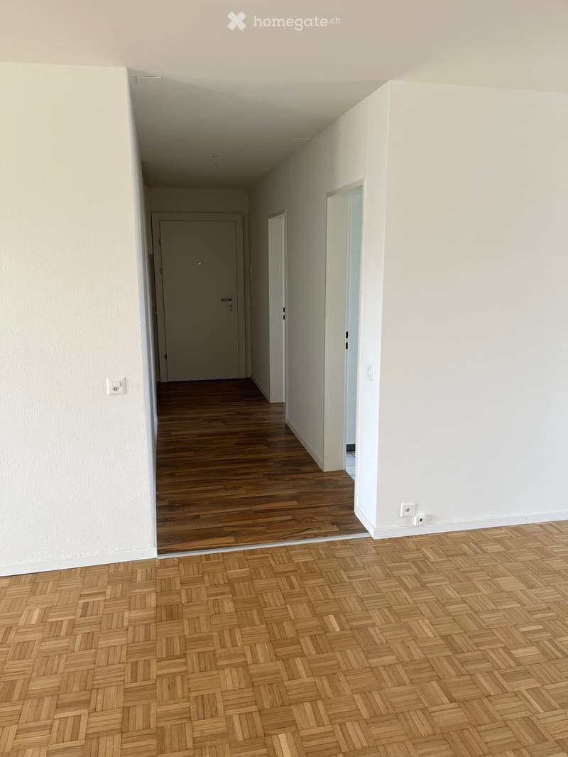 Empty hallway with light wooden flooring and white walls, featuring a door at the end and an open doorway leading to another room.