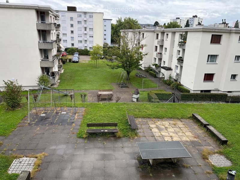 Courtyard with green lawns, benches, playground equipment, and a ping pong table, surrounded by apartment buildings.