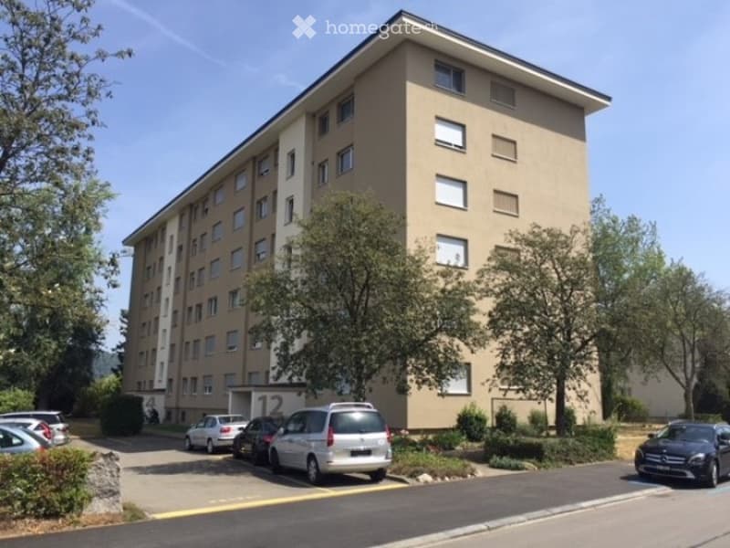 Multi-story residential building with numerous windows, surrounded by trees and parked cars.