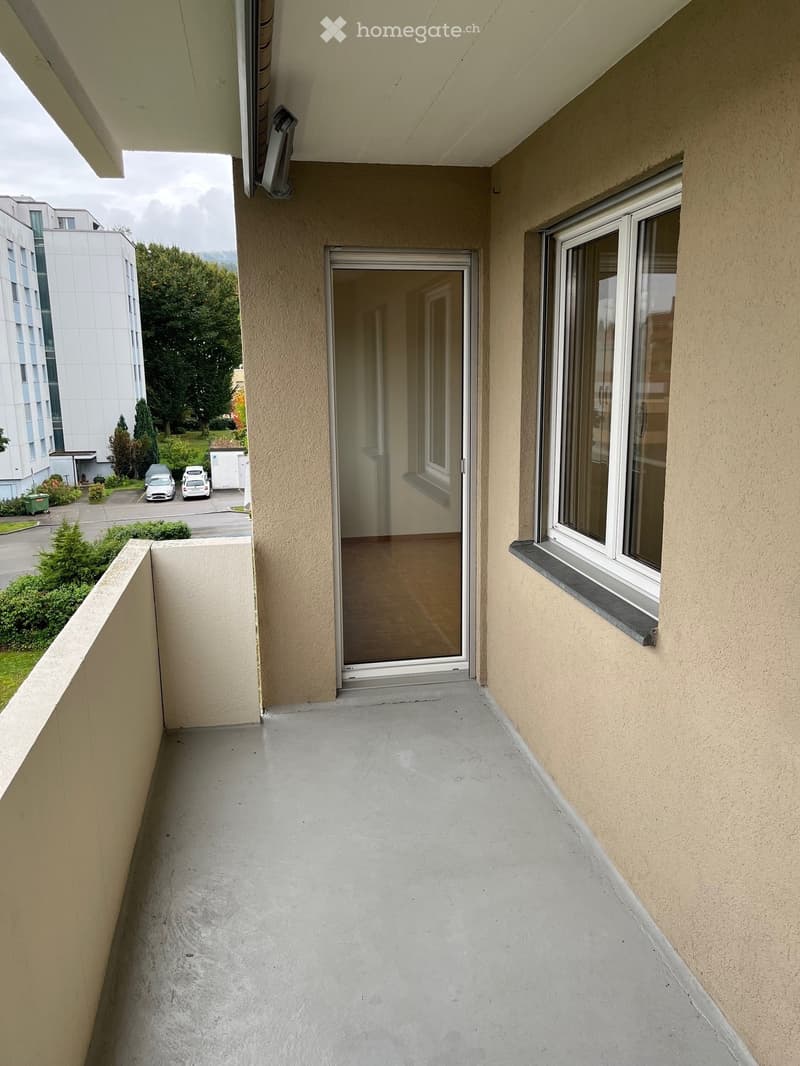 Empty balcony with concrete floor, glass sliding door, and window overlooking a parking area and greenery.