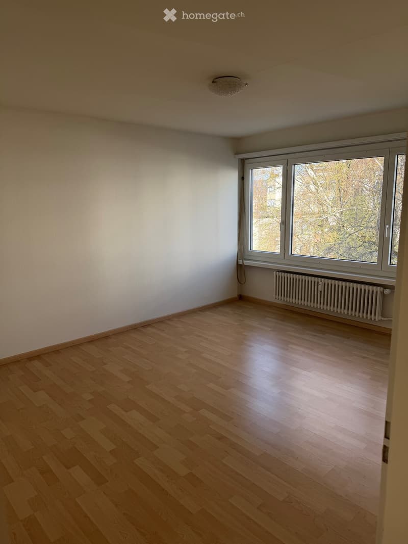 Empty room with wooden flooring, white walls, large windows, and a radiator below the windows.