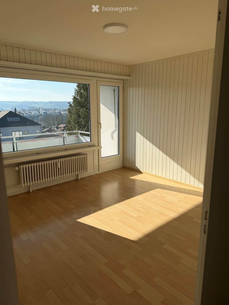 Empty room with wooden flooring, white paneled walls, large window with view, and a door leading to a balcony.