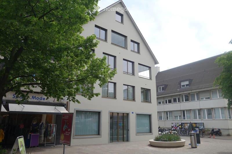 A modern office building with multiple glass windows, a white exterior, and a shop on the ground floor. There are bicycles parked to the right of the building, and a tree in front.