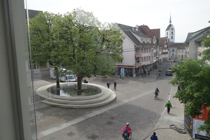 Aerial view of a city square with trees and people, central fountain, buildings on the sides, cars driving on the street