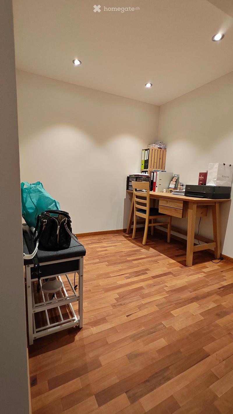 A small, well-lit room with wooden flooring, a desk with a chair, a printer, and a handbag on a stool.