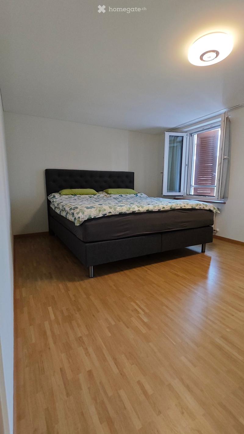 Bright bedroom with wooden flooring, a large bed with a dark headboard and green pillows, and a round ceiling light. The room has large windows with blinds.