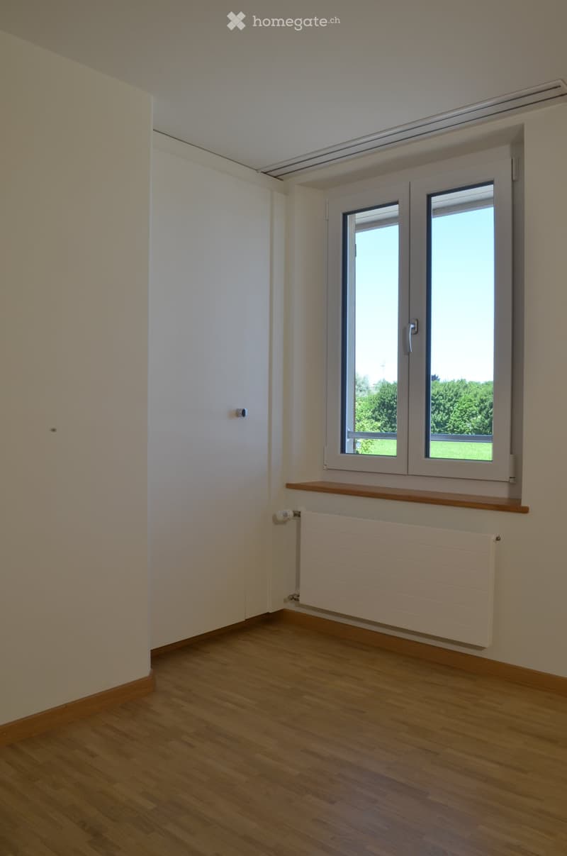 Empty room with wooden flooring, white walls, a window with a wooden sill, and a radiator below the window.