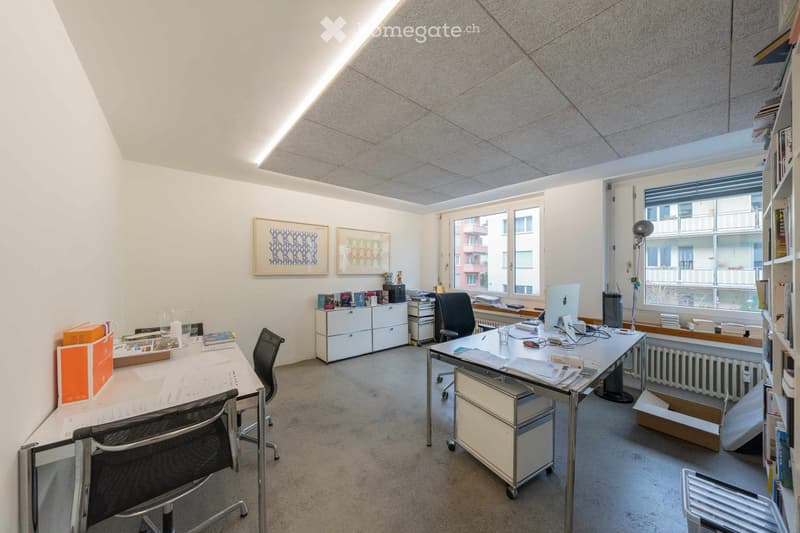 A spacious office room with desks, chairs, artwork, shelves, a radiator, and windows.